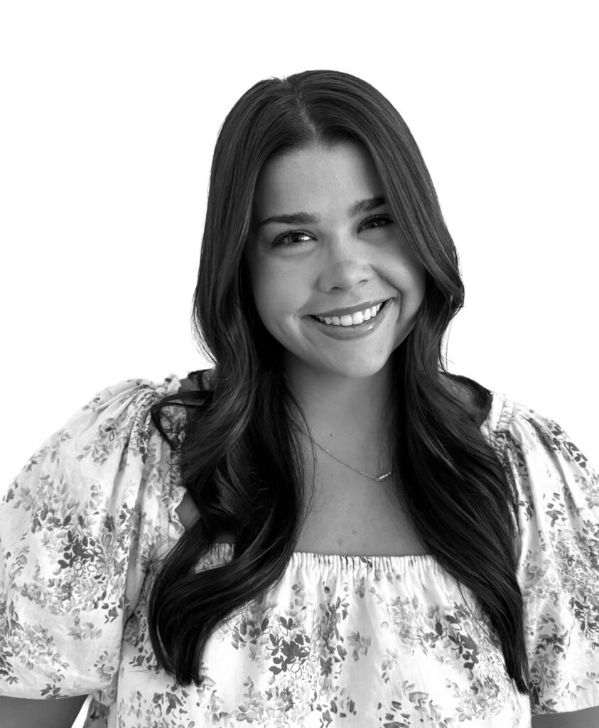 Black and white image of a person with long, dark hair, wearing a floral-patterned blouse and smiling about something at the camera.